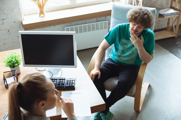 Young man sitting in office during the job interview with female employee, boss or HR-manager, talking, thinking, looks confident