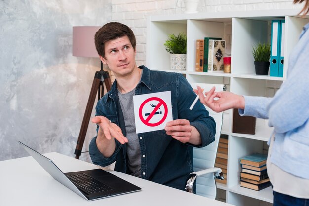 Young man sitting in office showing no smoking sign to woman holding cigarette