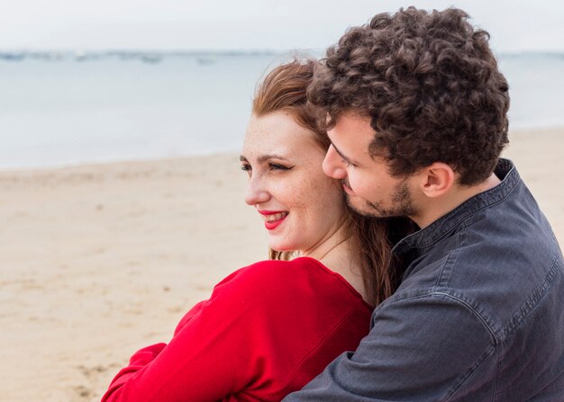 Молодой человек сидит на берегу моря и обнимает женщину