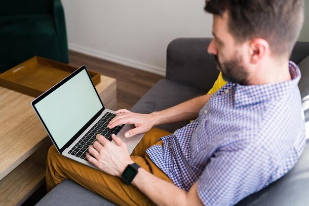 Young man sitting on the sofa and typing on his laptop with a blank screen and copy space