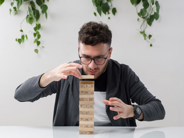 young man sitting in spectacles watches grey jacket playing with wooden figures along with plant on white