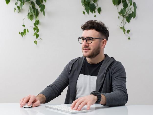 young man sitting in spectacles watches grey jacket working on pc along with plant on white