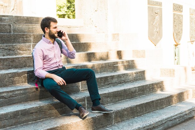 Молодой человек сидит на ступеньках и разговаривает по мобильному телефону