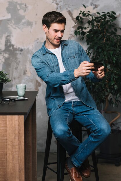 Молодой человек, сидя на стуле, берущий себя через мобильный телефон