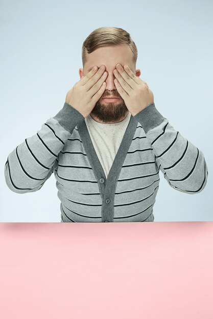 Young man sitting at table at studio with eyes closed isolated on blue.