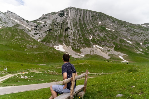 木製のベンチに座って、芝生のフィールドで山々の景色を楽しむ若い男