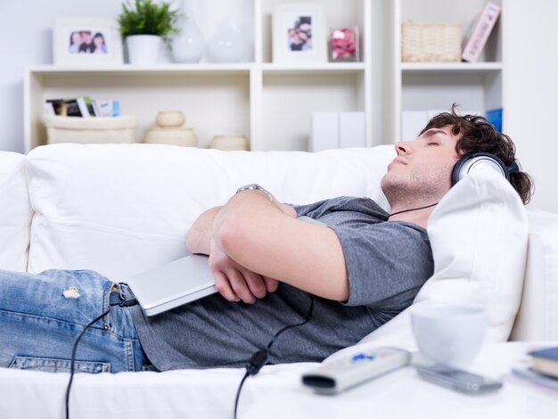 Молодой человек спит на диване дома с ноутбуком - в помещении