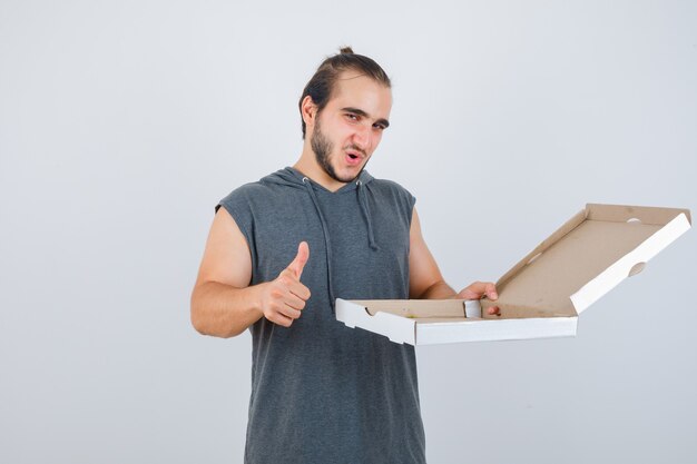 Young man in sleeveless hoodie holding opened pizza box while showing thumb up and looking delighted , front view.