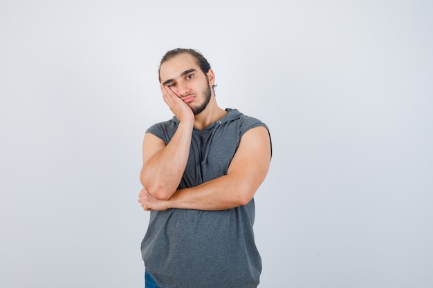 Young man in sleeveless hoodie keeping hand on cheek and looking thoughtful , front view.