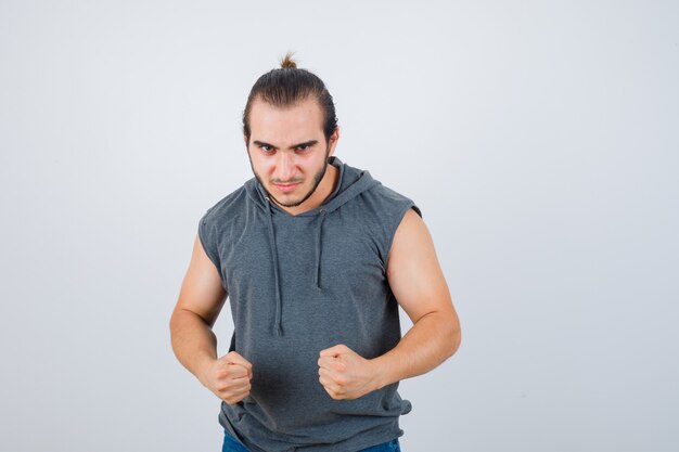 Young man in sleeveless hoodie standing in fight pose and looking spiteful , front view.