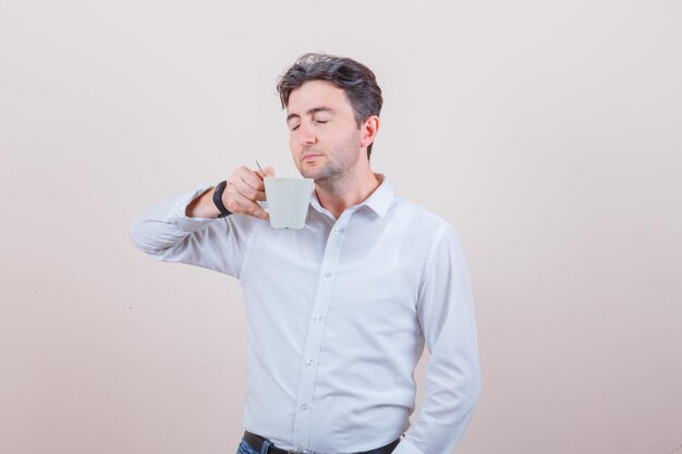 Young man smelling aromatic tea in white shirt, jeans and looking delighted