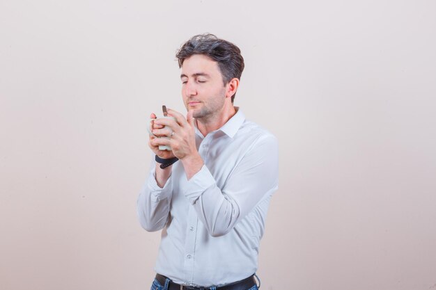 Young man smelling aromatic tea in white shirt, jeans and looking delighted