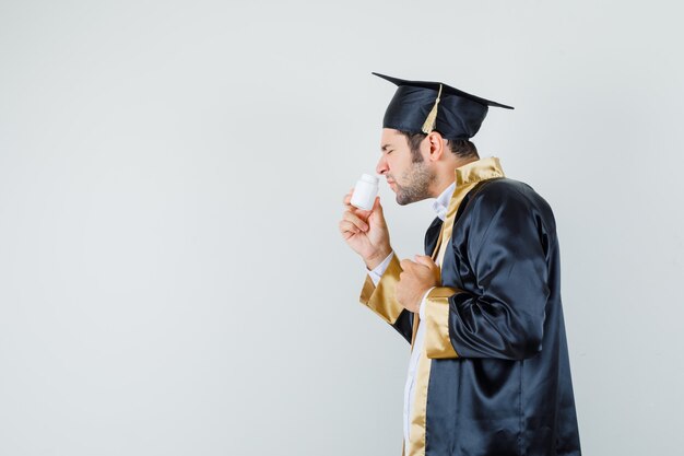 若い男は、卒業生の制服を着た瓶の中の錠剤の匂いを嗅ぎ、うんざりしているように見えます。