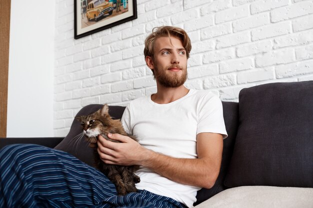 Молодой человек, улыбаясь, поглаживая кошку, сидя на диване у себя дома.