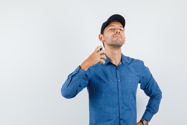 Young man spraying perfume on himself in blue shirt, cap and looking elegant , front view.