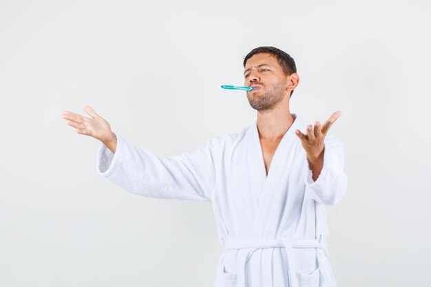 Young man spreading arms with toothbrush in white bathrobe and looking dissatisfied. front view.