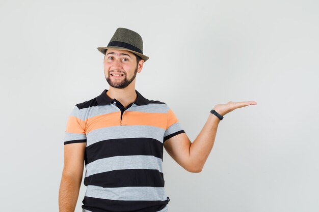 Young man spreading palm aside in t-shirt, hat and looking cheerful