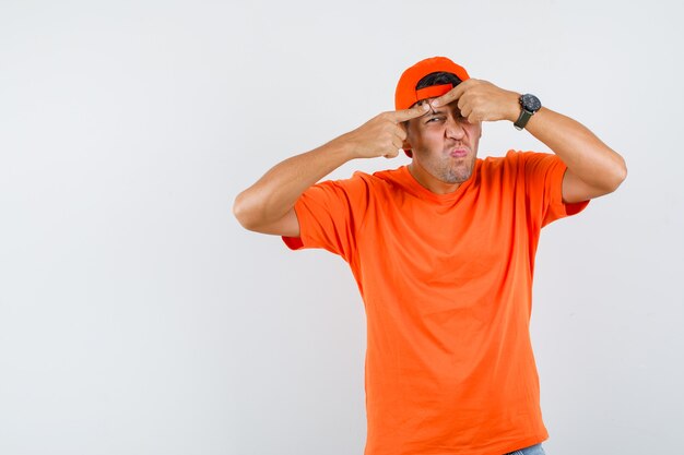 Молодой человек сжимает прыщ на лбу в оранжевой футболке и кепке, вид спереди.