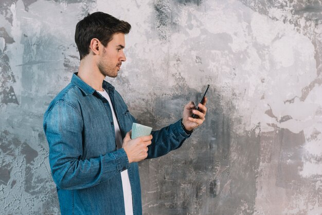 コーヒーカップを持っている携帯電話を見て風通しの壁の前に立っている若い男