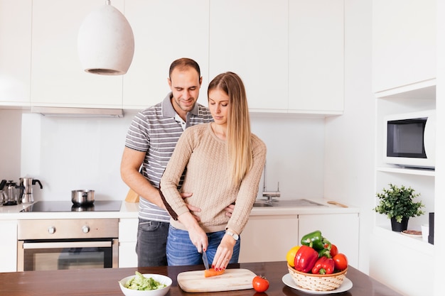 Free photo young man standing behind his wife cutting the carrot with knife on chopping board