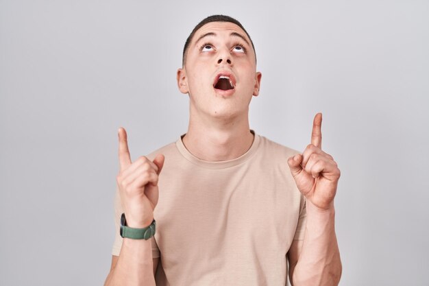 Young man standing over isolated background amazed and surprised looking up and pointing with fingers and raised arms