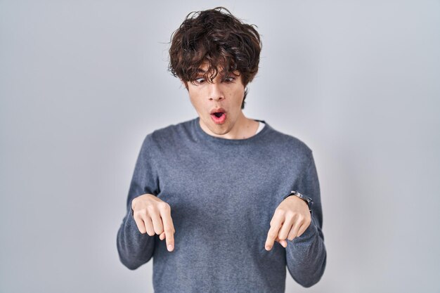 Молодой человек стоит на изолированном фоне, указывая пальцами вниз, показывая рекламу, удивленное лицо и открытый рот
