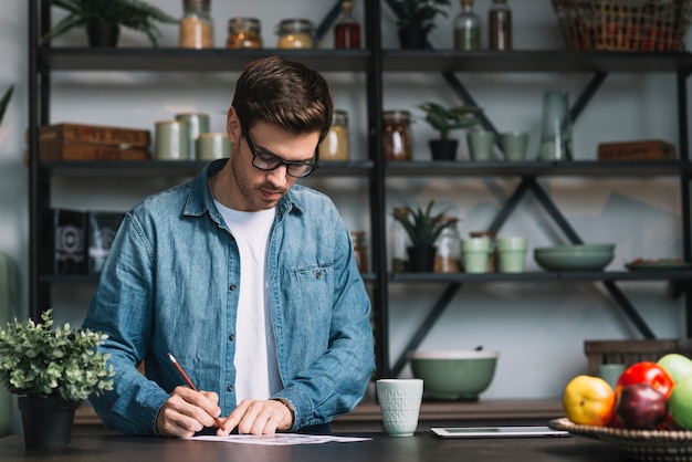 Young man standing behind the kitchen counter writing on paper with pencil