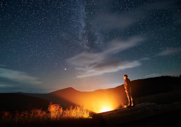 Young man standing on logs near campfire in the mountains under sky full of stars