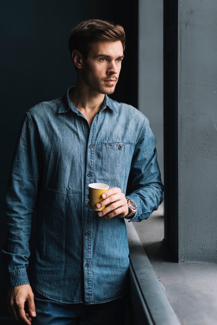 Free Photo young man standing near the window holding disposable coffee cup