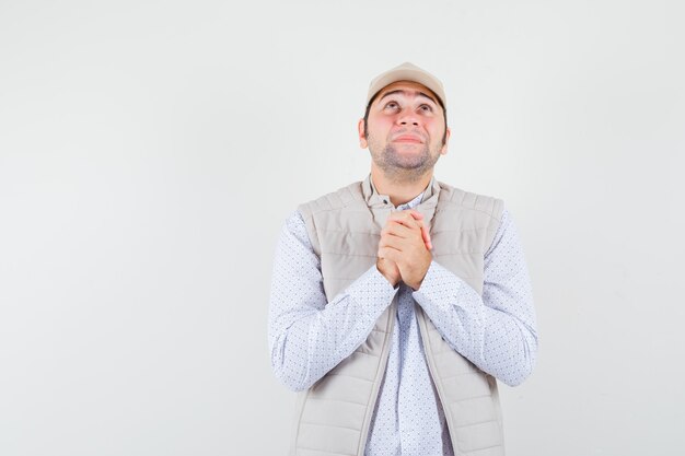 Young man standing in prayer pose and looking upward in beige jacket and cap and looking focused. front view.