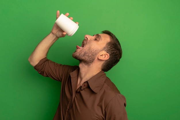 Young man standing in profile view holding plastic coffee cup above head looking inside it trying to drink coffee isolated on green wall