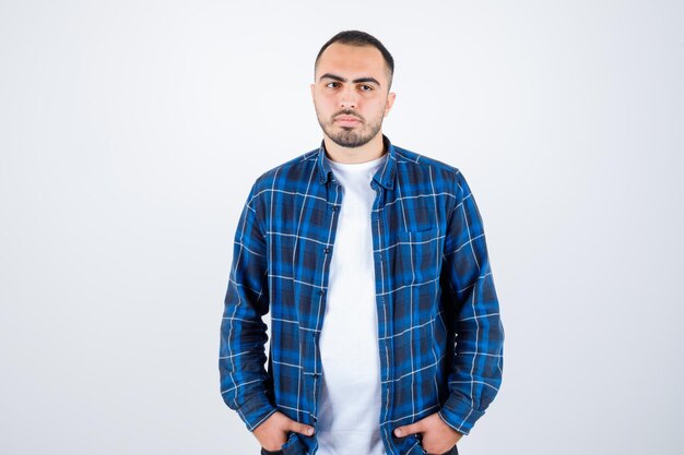 Young man standing straight and holding hands in pocket in checked shirt and white t-shirt and looking serious