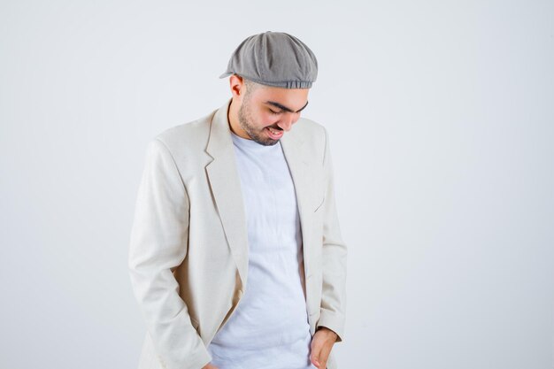 Young man standing straight, looking down and posing at front in white t-shirt, jacket and gray cap and looking happy