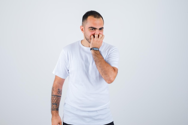 Young man standing straight, putting hand on mouth in white t-shirt and black pants and looking surprised