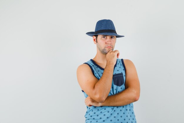 Young man standing in thinking pose in blue singlet, hat and looking serious. front view.