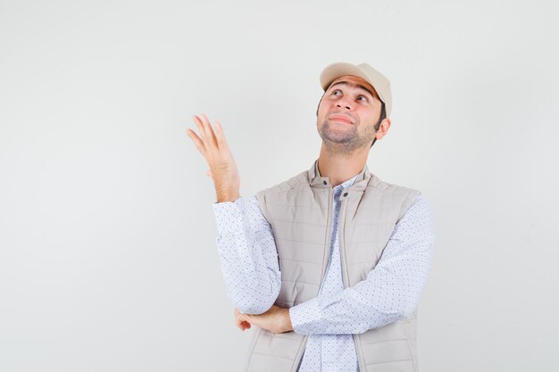 Young man standing in thinking pose, looking away in beige jacket and cap and looking happy , front view.