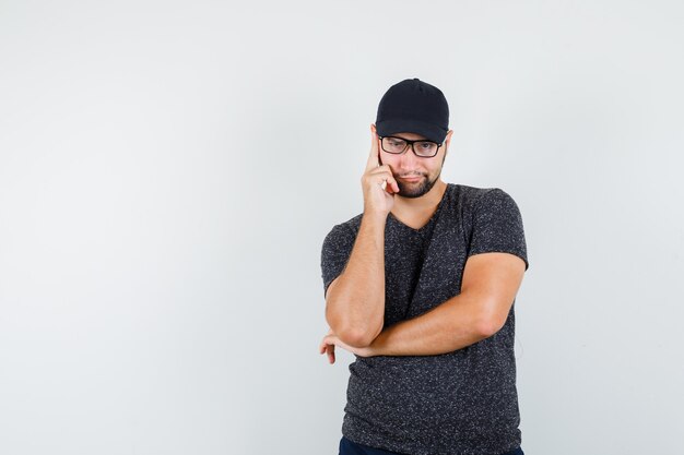 Young man standing in thinking pose in t-shirt and cap, jeans and looking sly