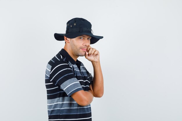 Young man standing in thinking pose in t-shirt, hat and looking jolly. front view.