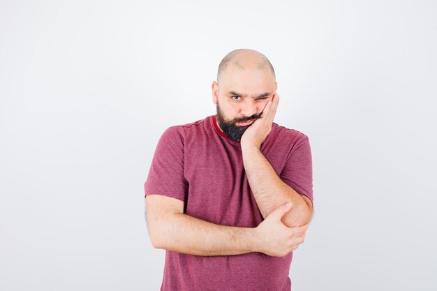 Young man standing in thinking pose while leaning cheek on palm in pink t-shirt and looking pensive , front view.