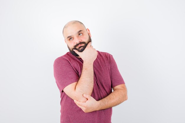 Young man standing in thinking pose while putting hand on chin in pink t-shirt and looking pensive. front view.