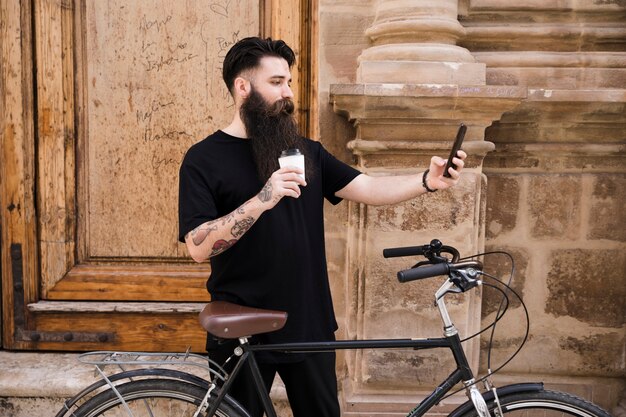 Молодой человек стоя с велосипедом перед деревянной дверью принимая selfie на мобильном телефоне