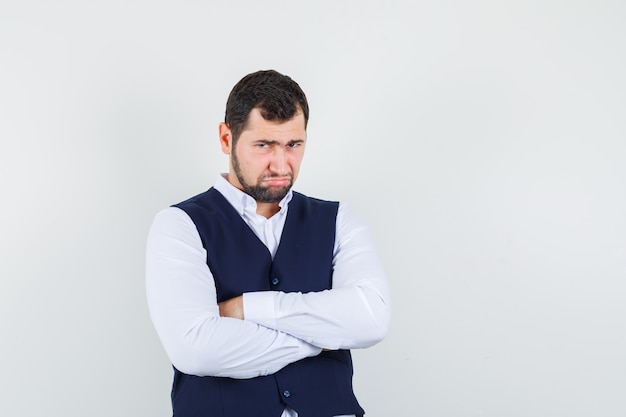 Young man standing with crossed arms in shirt and vest and looking offended