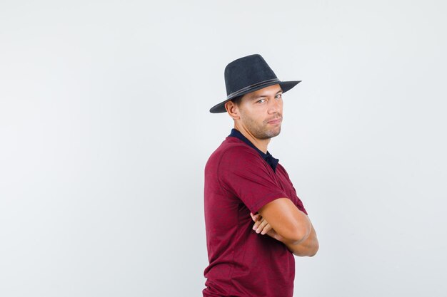 Young man standing with crossed arms in t-shirt, hat and looking optimistic .