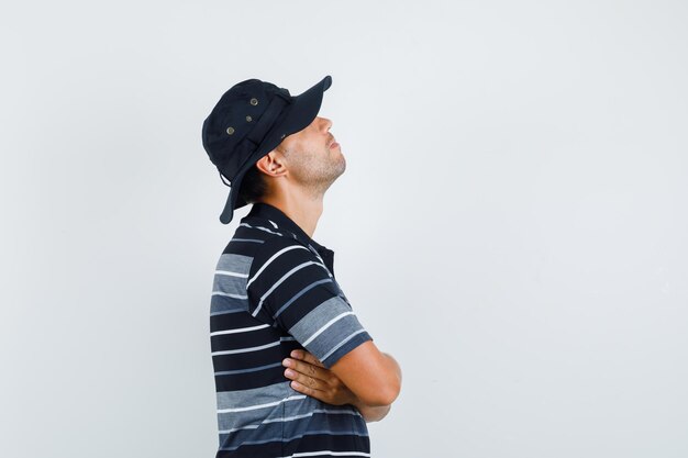 Молодой человек, стоящий со скрещенными руками в футболке, шляпе и задумчивый.