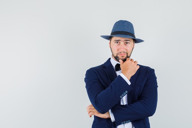 Giovane uomo in piedi con il dito sul mento in tuta, cappello e guardando fiducioso, vista frontale.