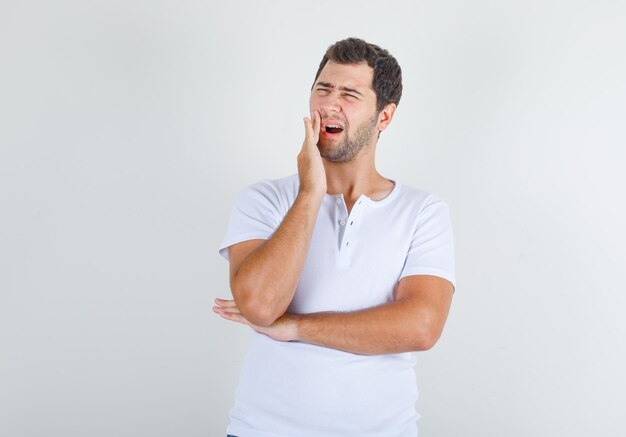 Young man standing with hand near mouth in white t-shirt and looking painful