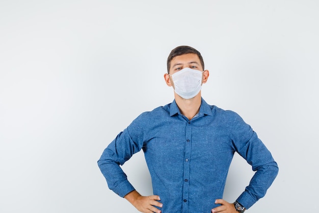 Young man standing with hands on waist in blue shirt, mask and looking serious. front view.