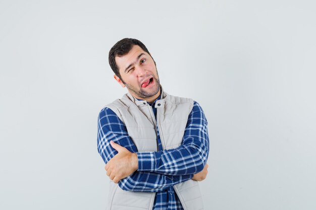 Young man sticking tongue out while winking in shirt,sleeveless jacket and looking funny , front view.