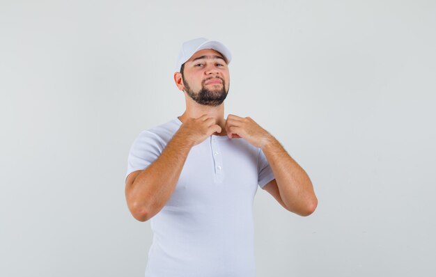 Young man straightening his collar in t-shirt,cap and looking ready. front view.
