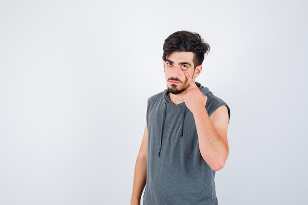 Young man stretching eye area with index finger in gray t-shirt and looking serious